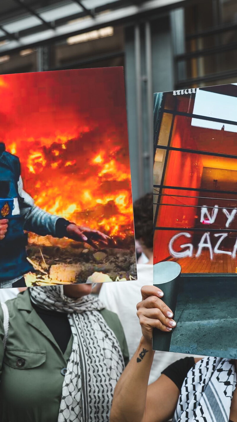 People hold up posters and signs at a protest. The poster in the center of the image is a photo of martyred Al Jazeera journalist Anas al-Sharif, killed by Israel on August 10, 2025. Next to it is a poster that reads: "NYT LIES, GAZA DIES"