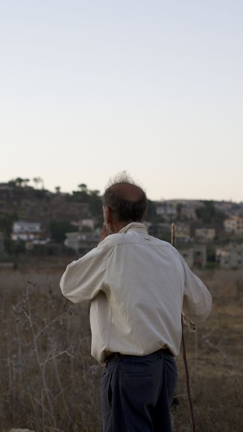 A man stands in a dry field looking toward a hillside town at dusk.