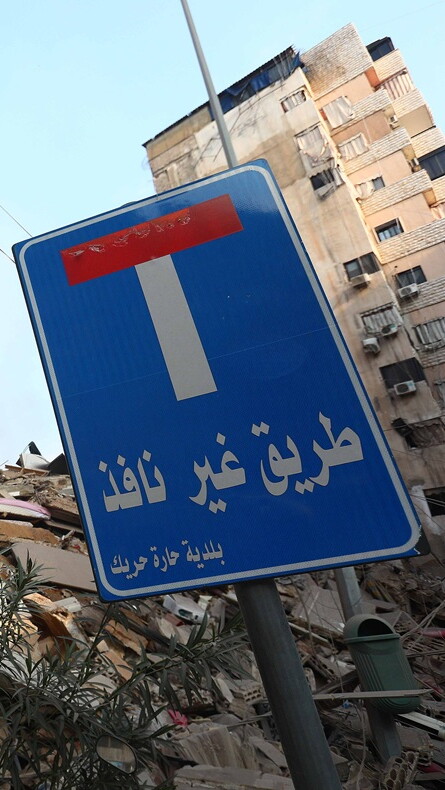 في وسط الصورة، يقف رجل فوق كومة من الأنقاض. إلى اليمين، في المقدمة، لافتة كُتب عليها: "طريق مسدود".
