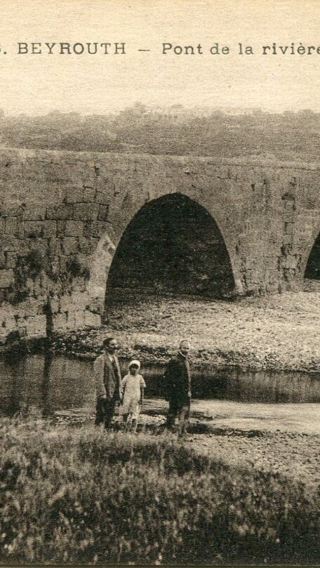 An arched stone bridge crosses the Beirut River; mountains are visible in the background. Three people are walking by the river. 