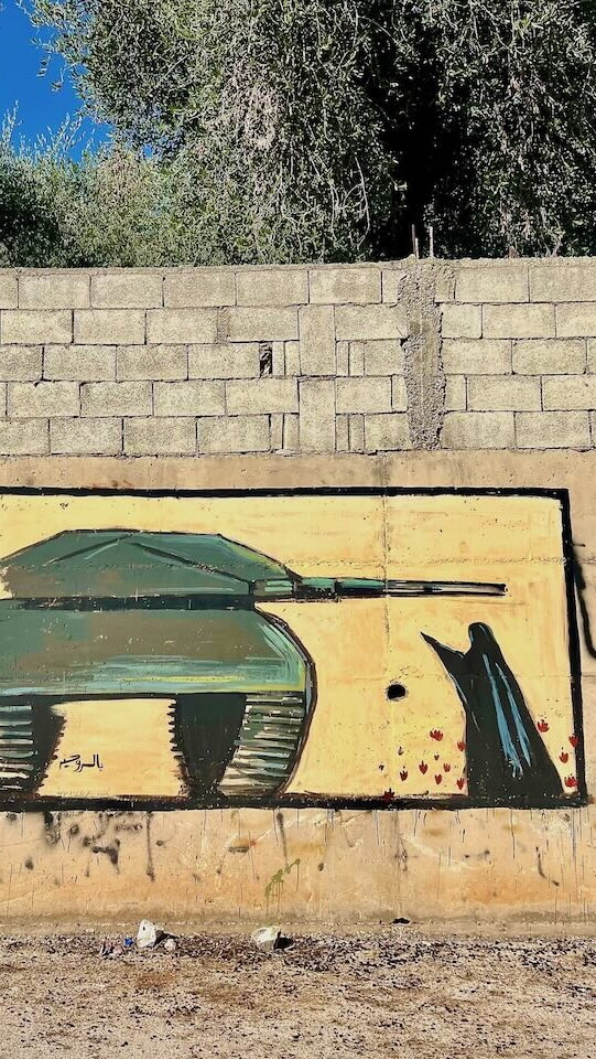 A mural of a green military tank is painted on a concrete wall with graffiti and a low brick barrier above; a woman is confronting the tank; trees and blue sky are visible in the background.
