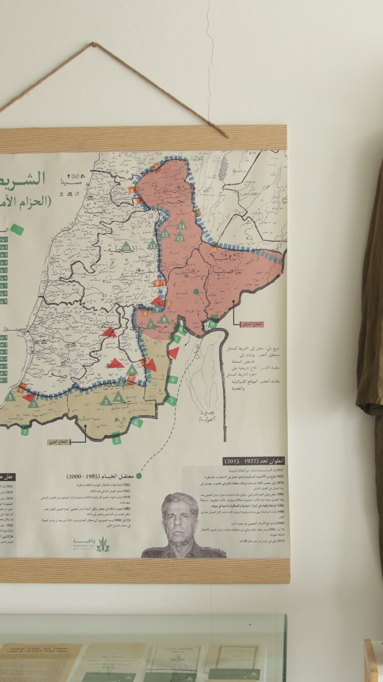 An exhibition wall featuring a large framed border map with Arabic text on the left, and a full-length olive-green military jumpsuit with a matching boonie hat hanging on the right.