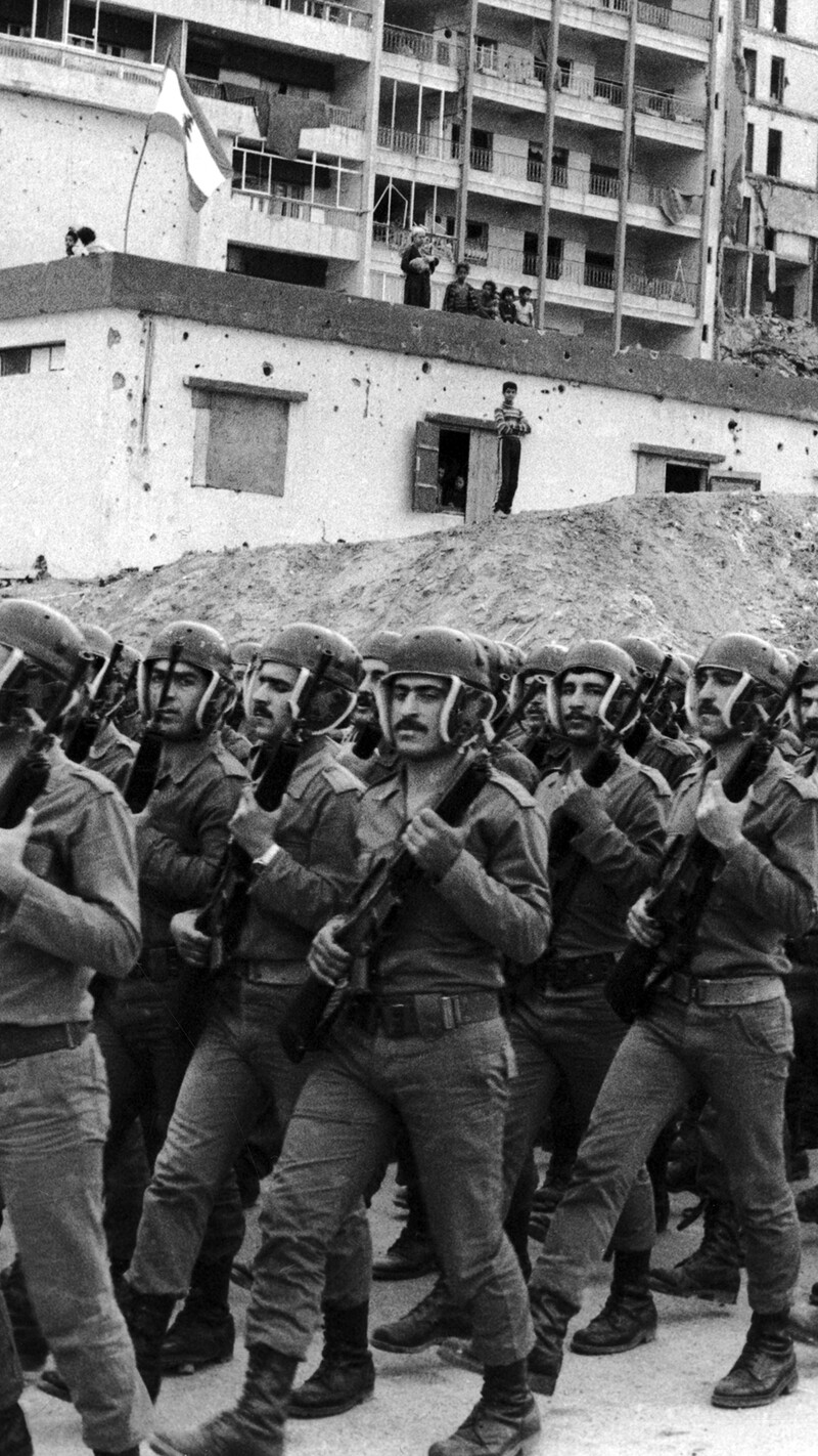 A black-and-white photograph of a large formation of Lebanese Army soldiers marching in tight unison during a drill. The soldiers are dressed in matching uniforms and combat boots, wearing distinctive helmets with integrated ear and chin protection, holding rifles diagonally across their chests. In the background, a war-torn urban landscape features large mounds of dirt and rubble in front of a heavily damaged, multi-story apartment complex and a smaller white building marked with bullet holes.