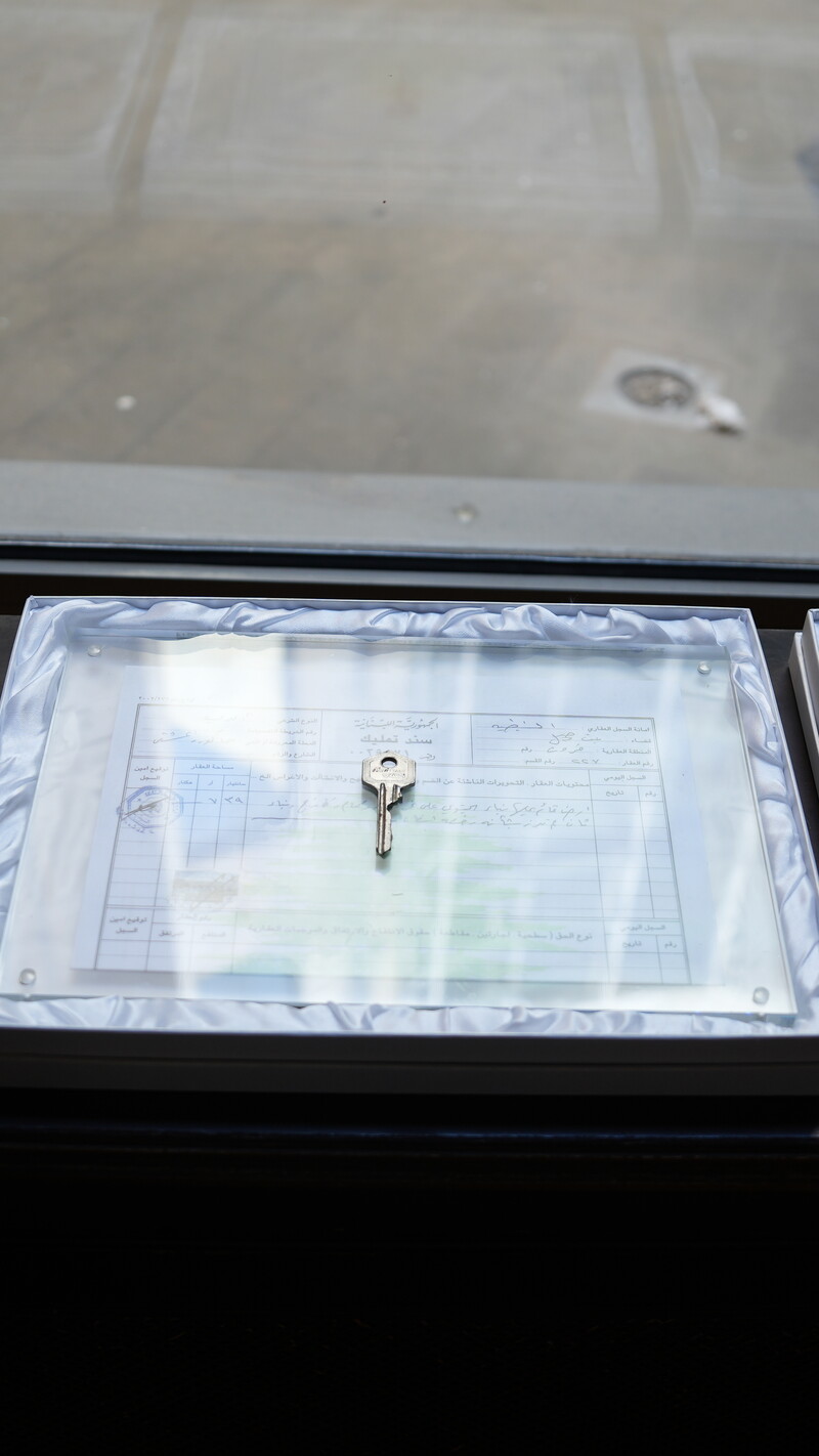 Three framed displays rest on a windowsill, each containing a key placed over a visual object: a photograph of a damaged house, a title deed, and an aerial image of a marked area. 