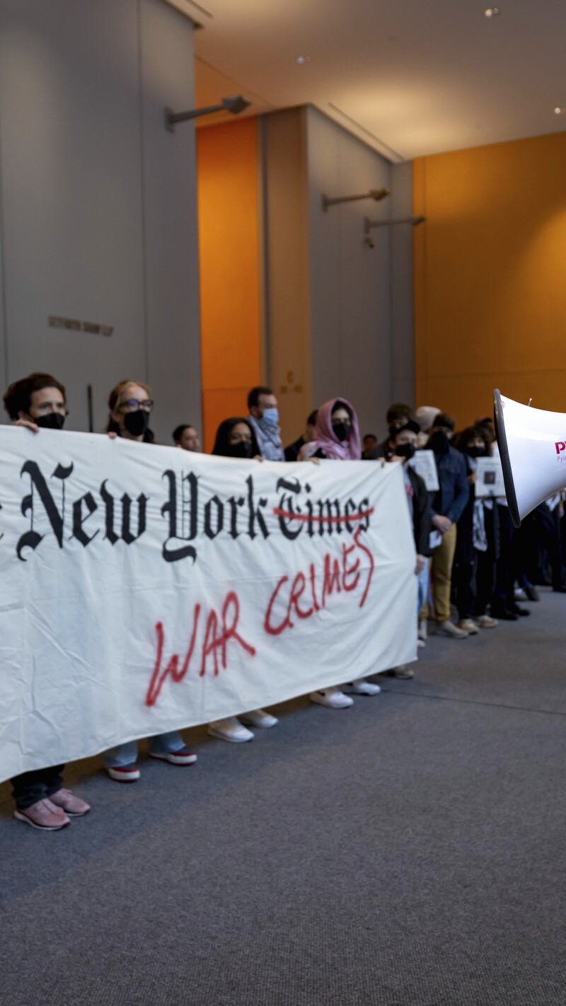 Protesters at a demonstration inside The New York Times building, holding a banner accusing the publication of coverage related to war crimes, with one person speaking through a megaphone.