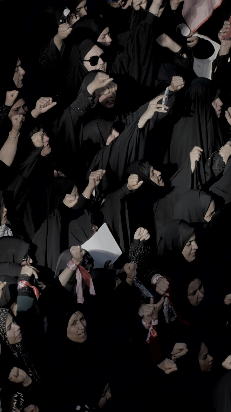 A high-angle view of a tightly packed crowd, largely composed of women in black chadors, raising their clenched fists together during a large gathering, with an Iranian flag visible among the group.