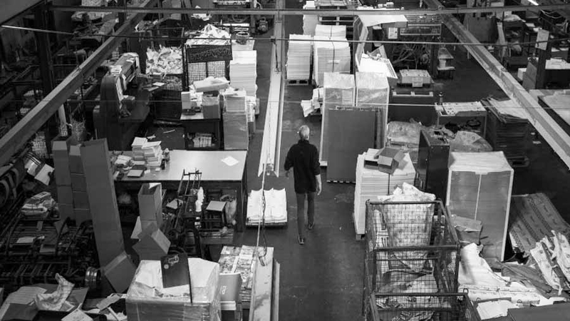 A person walks through a warehouse filled with stacks of paper, boxes, and various materials.