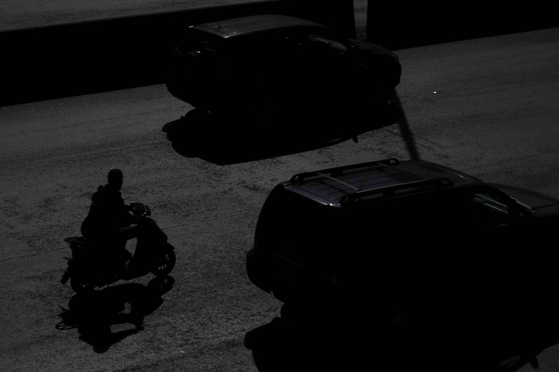 Dark photo of delivery worker driving motorcycle on a road behind two cars.