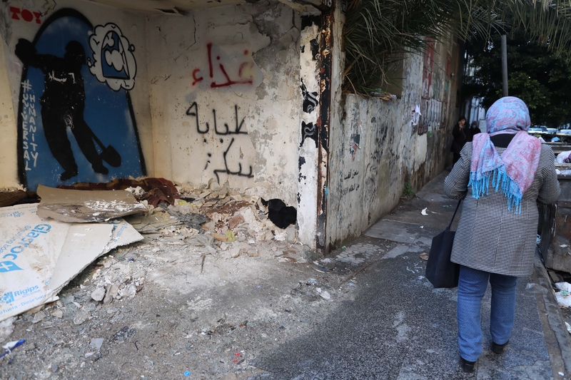 A caved-in room in Hamra, decorated with graffiti -- its walls read: (left) "Universal humanity"; (right) "Nourishment, healthcare, housing." The collapsed wall gives way to a thick blanket on the ground, seemingly left behind.