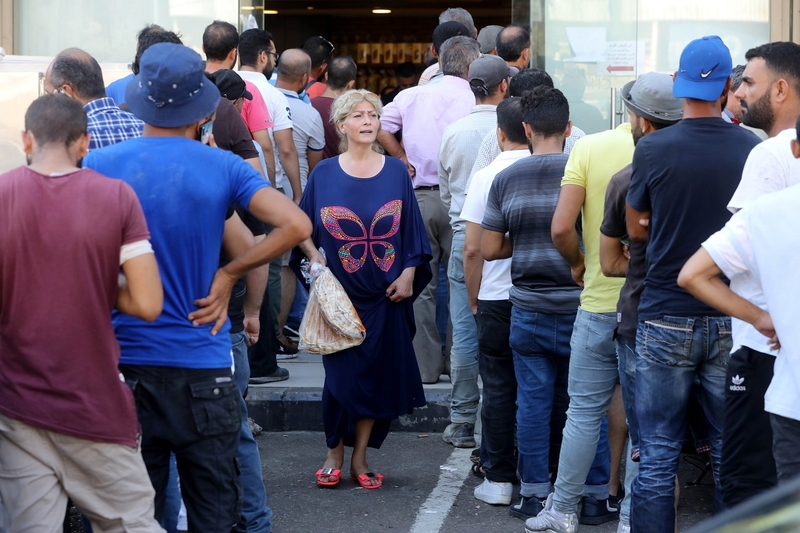 Facing the camera, a woman in a colorful dress and pink slipper shoes walks out of a bakery holding a bag of Arabic bread, between two rows of men turned away from the camera, queuing.
