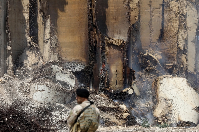 A man in a Lebanese Armed Forces camo uniform and black beret stands with back to the camera in front of a smoldering structure.