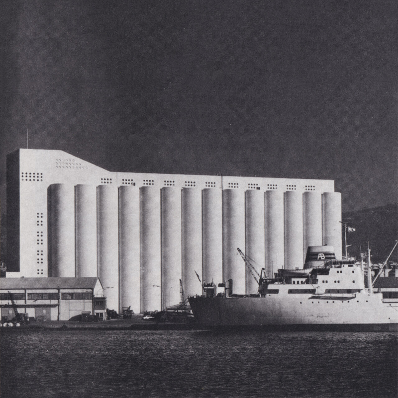 A black and white image of the Beirut grain silos which consist of a white concrete building with 14 cylinder shapes. The sea and a boat can be seen in the foreground.