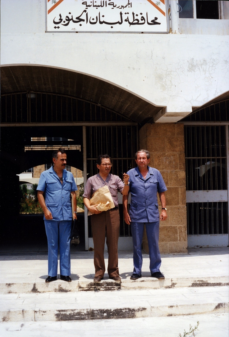 Three men stand outside of a heavily-gated building. The men on the left and right are dressed fully in blue.