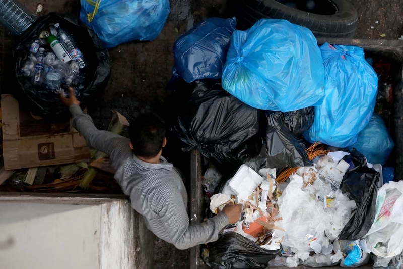 A waste picker is seen from above sorting through a garbage bin. He is collecting plastic bottles in a bag.