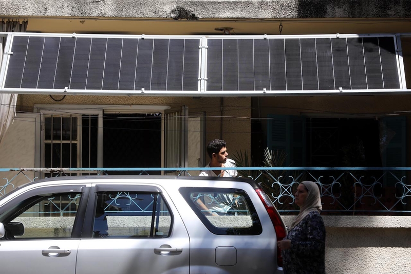 A man stands on a ground-floor balcony adjacent to the road, as solar panels hang above him. Only a few meters in front, a woman walks by a parked car.