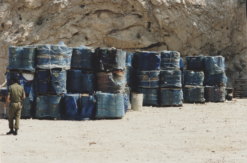 Barrels of toxic waste at a quarry. A uniformed man in green and in boots stands to the left of them.