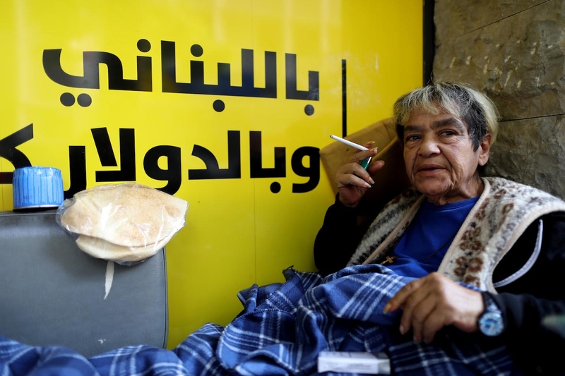 An old woman smoking a cigarette sits in from an OMT signs that reads: “In Lebanese liras and dollars too.” 