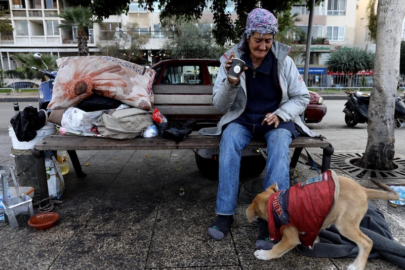 An old woman sits on a bench next to her belongings. She is holding a paper cup and looking at a puppy which is wearing a red jacket.