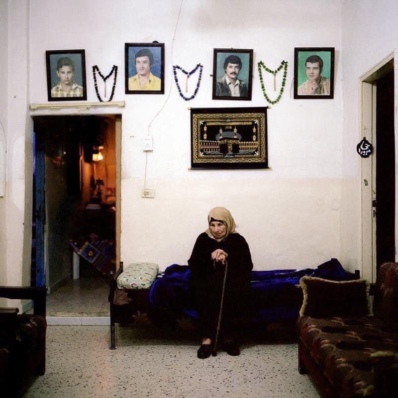 A woman sits on a single bed, placed against a living room wall with four portraits hanging and worry beads between each portrait. A tapestry of the Kaaba hangs underneath the portraits.