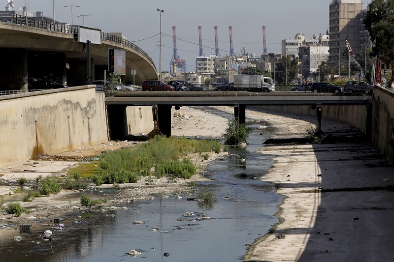 A meager amount of water passing through a mostly-empty concrete canal in a dense city. A bridge with traffic crosses over. A port looms on the horizon. 