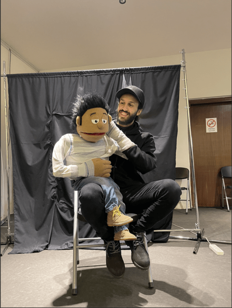 A smiling young man dressed in black is sitting on a chair and holding up a puppet.