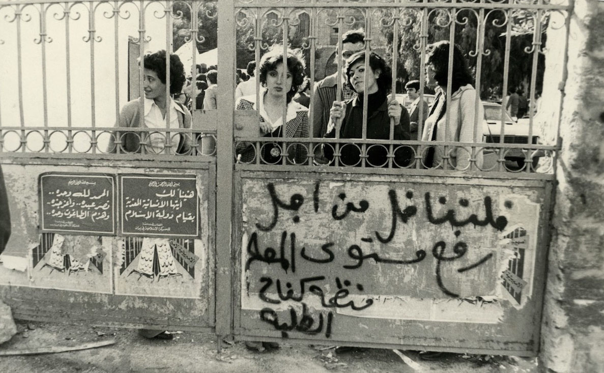 A group of students stand behind a gate, the right half of which is tagged: “Let us fight to raise the level of education (or of the teacher),” signed, “The Organization for Student Struggle.” On the left half of the gate are two posters signed, “The Committees in Solidarity with the Islamic Movement in Iran.” The first poster reads: “Congratulations to you, oh tortured humanity, for the rise of the Islamic State.” The second contains a quote from Takbeerat Al-Adha: “Praise be to God alone.. Gave 