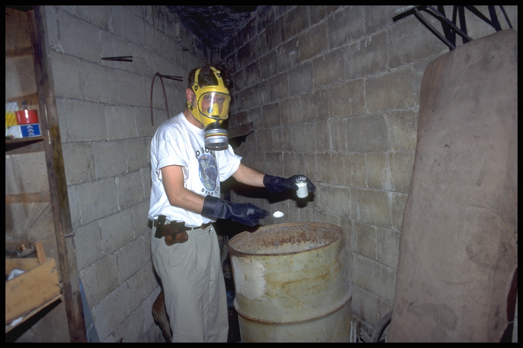 Man wearing gasmask and gloves holding jar and sample in spoon. He is standing by a barrel. 