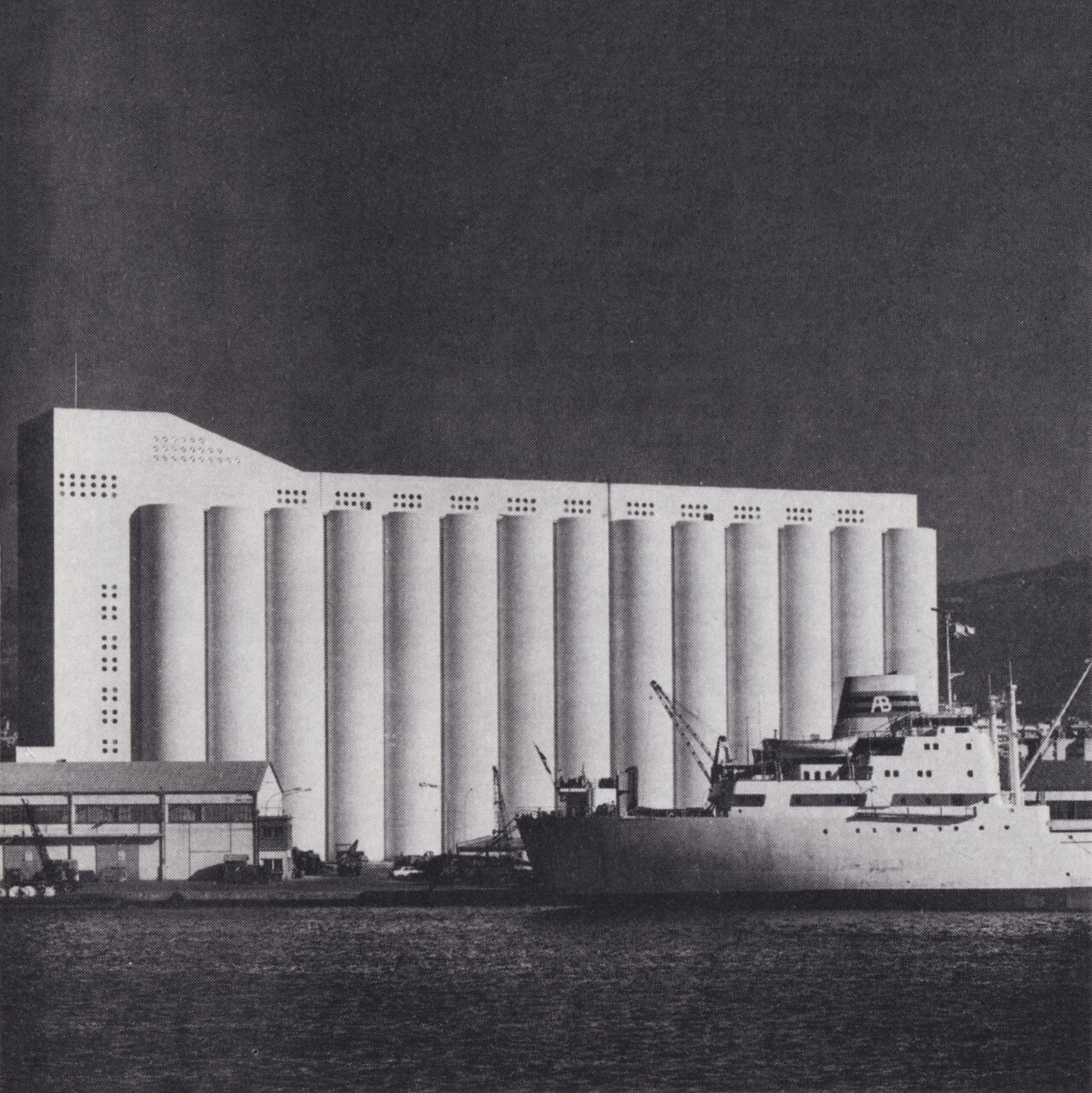 A black and white image of the Beirut grain silos which consist of a white concrete building with 14 cylinder shapes. The sea and a boat can be seen in the foreground.