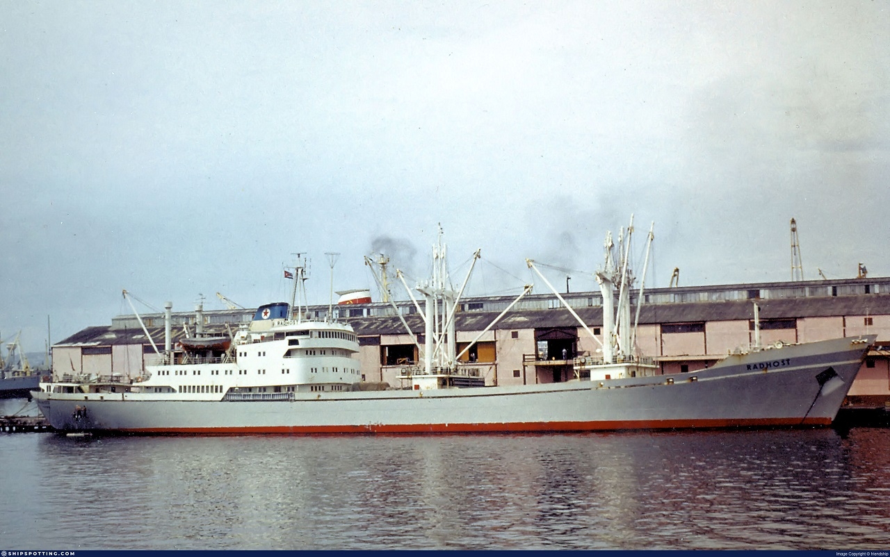 Cargo ship sits on the water, docked at a port. There is a warehouse in the background and slight smoke in the air.