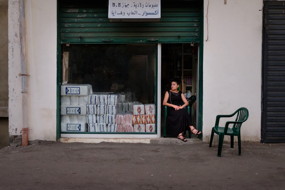 A woman sits on a plastic chair at the entrance of a small store. It is pitch black inside.