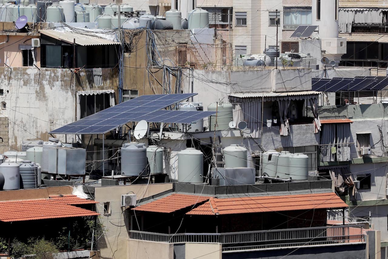 Crowded rooftops show solar panels amid water tanks and satellite dishes. 