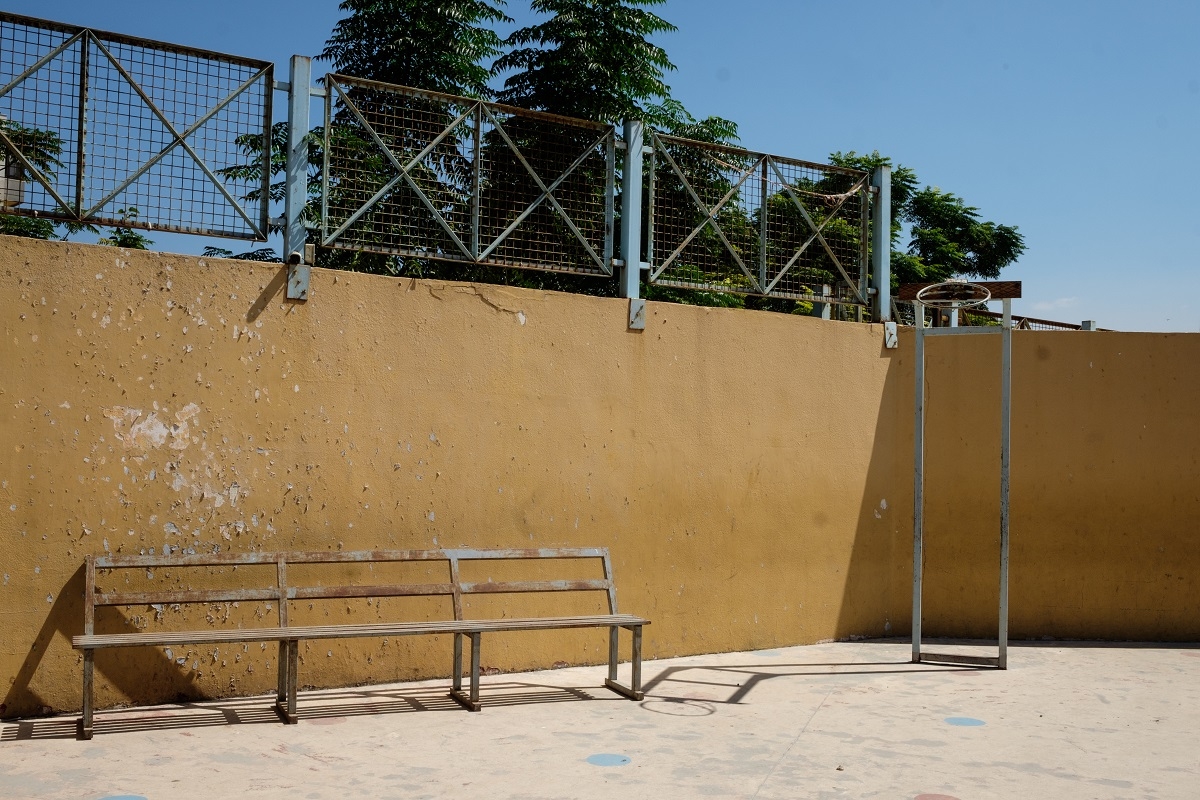 A now-rusty, once-blue bench in front of a yellow wall in the courtyard of a school. The yellow paint is peeling off the wall. To the right of the bench is a similarly rusty basketball hoop short of a net.