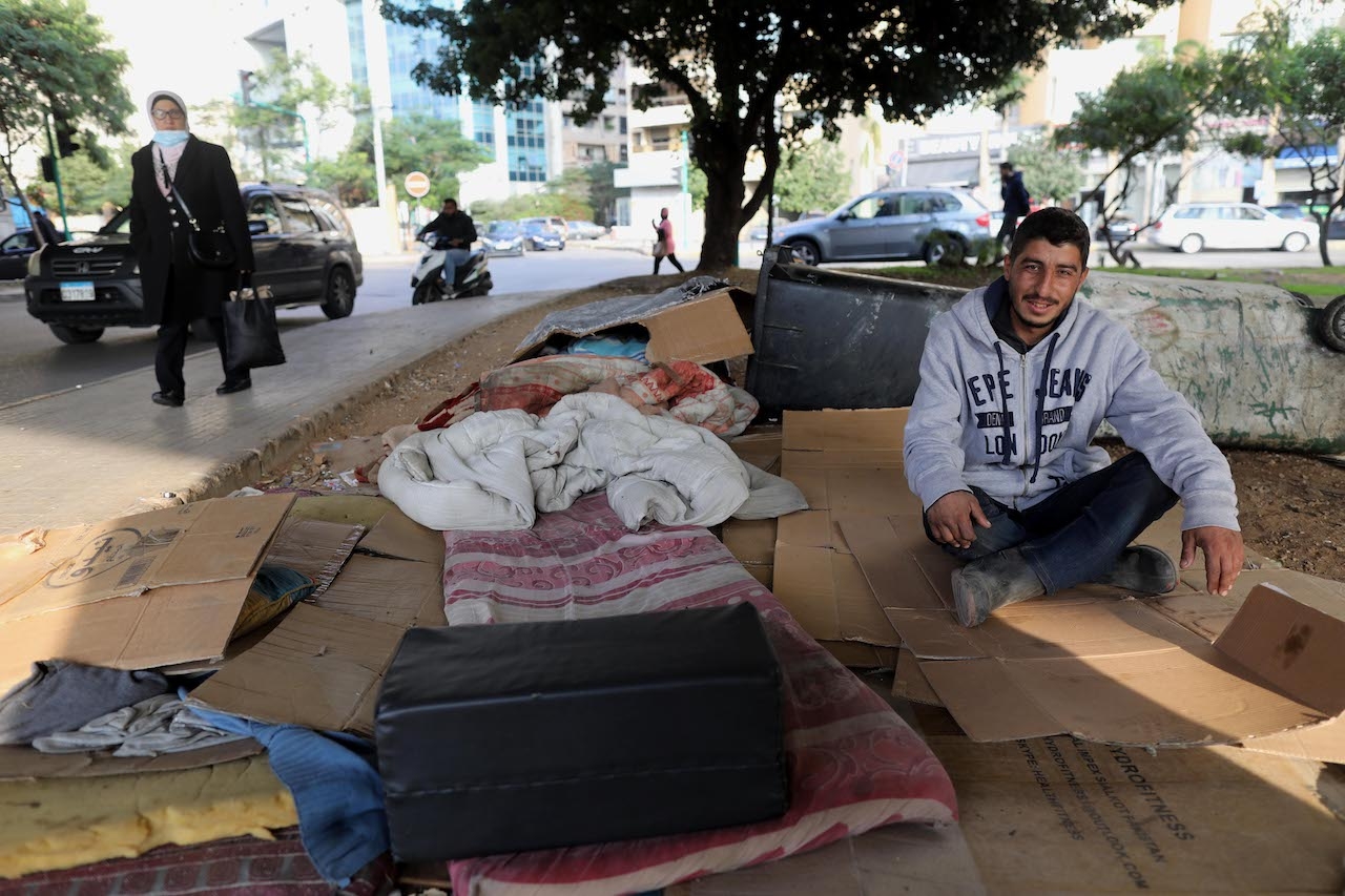 A young man sits on cardboards in the street next to blankets and his belongings.