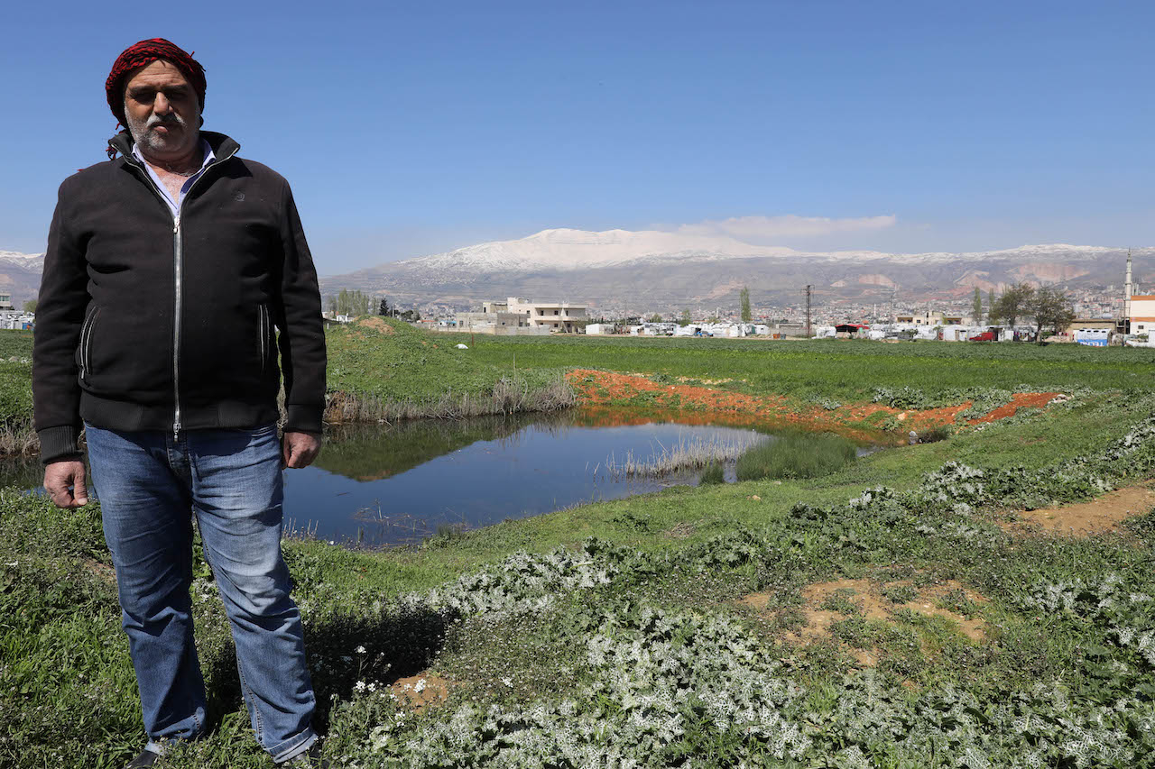 A farmer wearing a black jacket, jeans, and a red kouffiyeh on his headd stands over a small water pond in the middle of an agricultural field. A snowy mountain range appears in the background