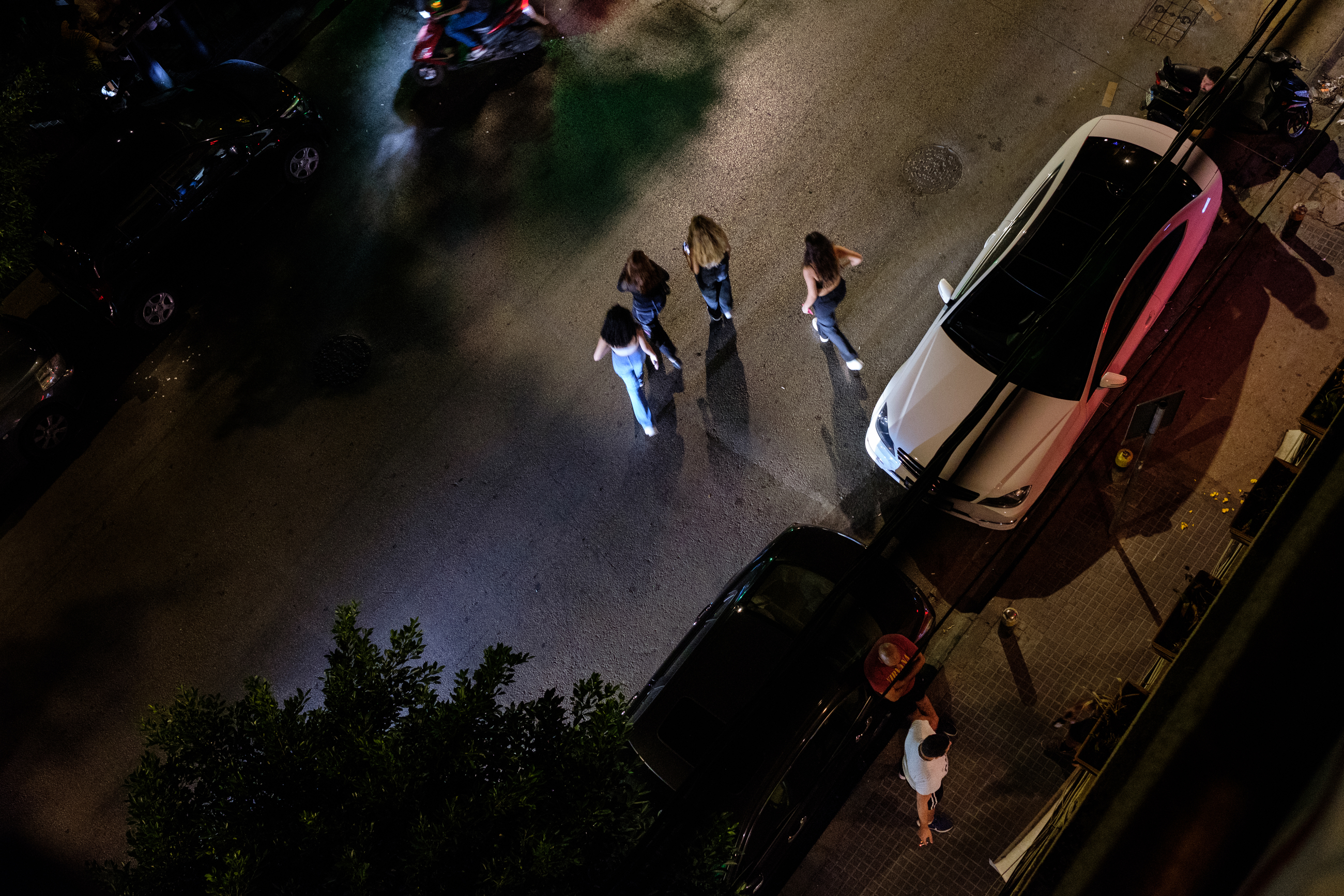 Four women cross the street of Gemmayze during the night, surrounded by cars parked on the side of the road.