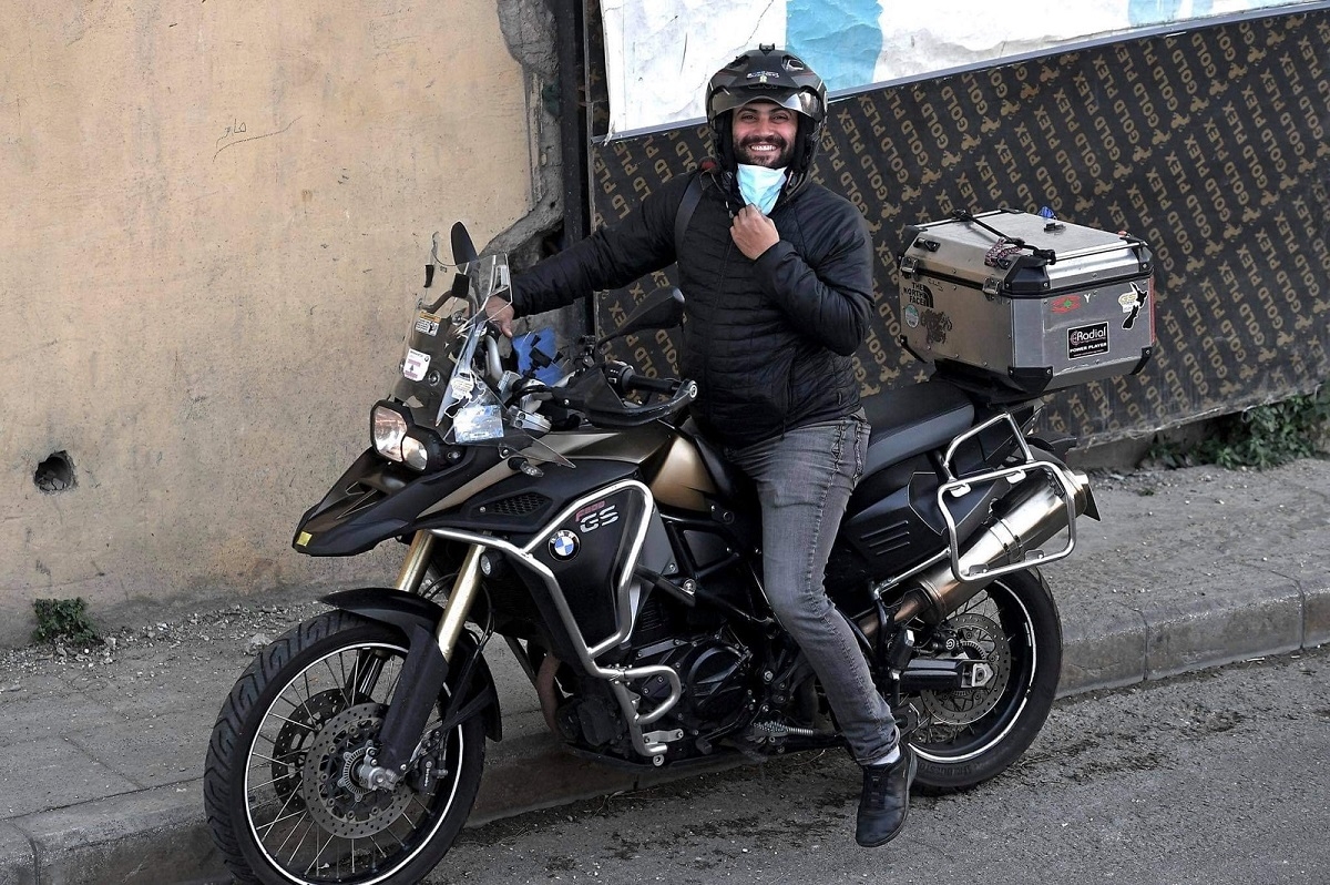 Helmet-adorned journalist Issam Abdallah pulls down a surgical mask and smiles on the back of a BMW F 800 GS Adventure motorcycle.