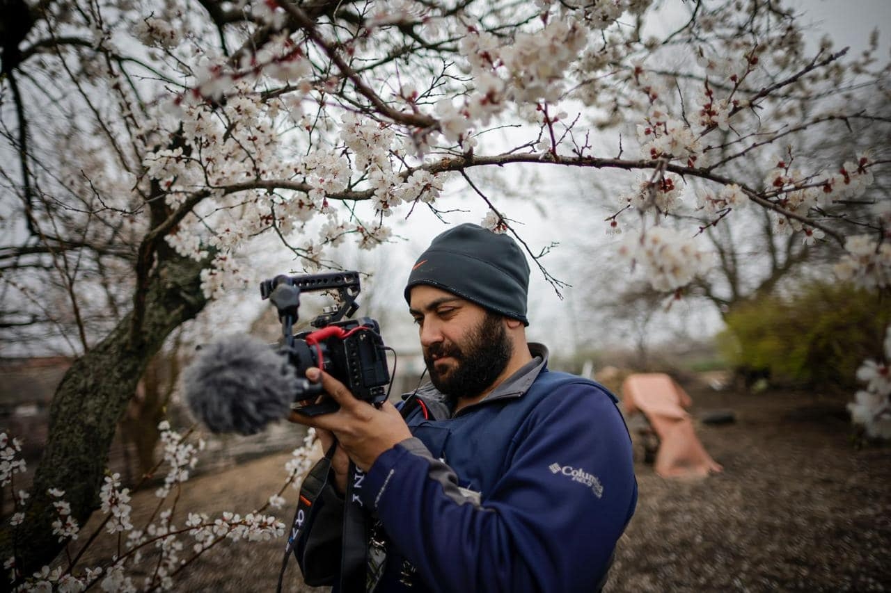 Journalist Issam Abdallah wears a beanie and holds a camera among flower blossoms and a grey winter sky.