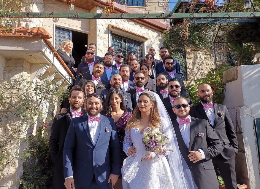 A large group of a couple of women and mostly men in suits wearing pink bowties surround a bride outside of a stone country home on a sunny winter day.