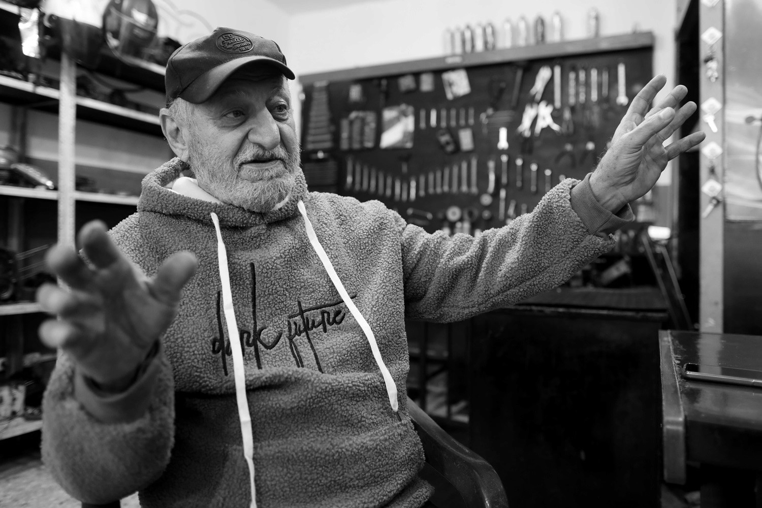 A man with a thick mustache wearing a baseball cap and hoodie speaks, and motions with his hands.