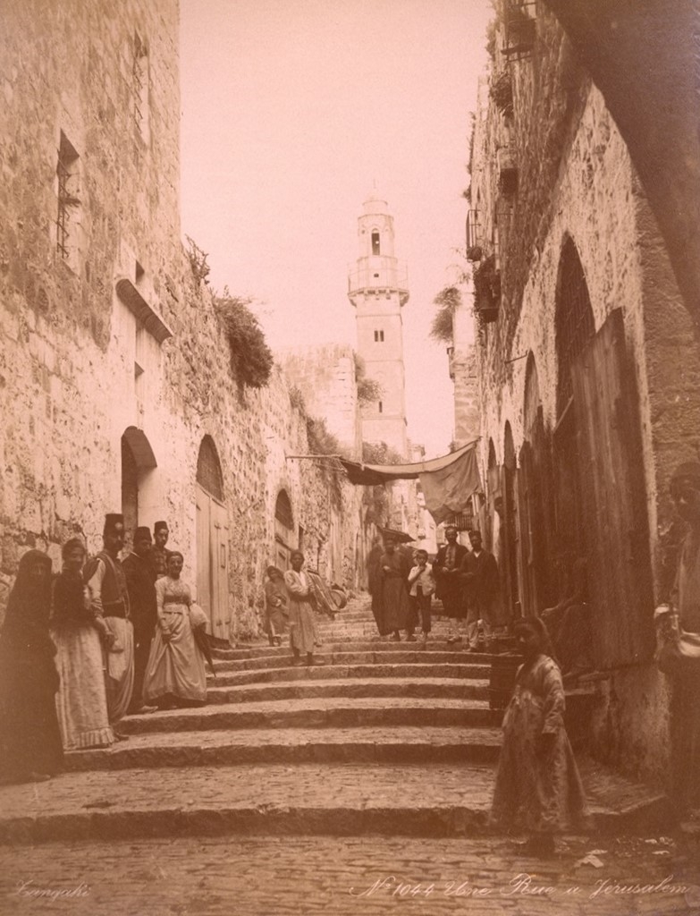 A sepia-colored photograph. A group of people strolling along a narrow street.