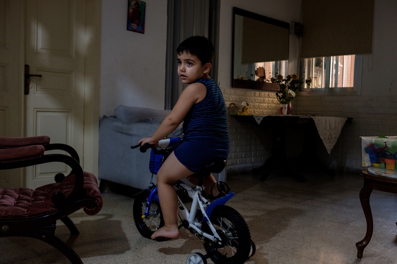 A little boy sits on a tricycle inside a dimly lit family home.