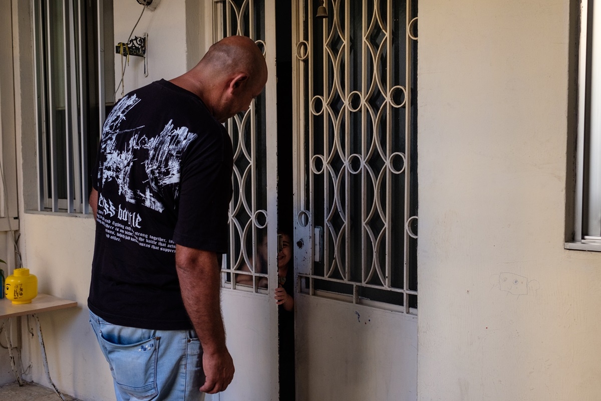 A tall man wearing a graphic t-shirt peers into the crack of front door, down at a little boy who is smiling and playing from the inside of the house.