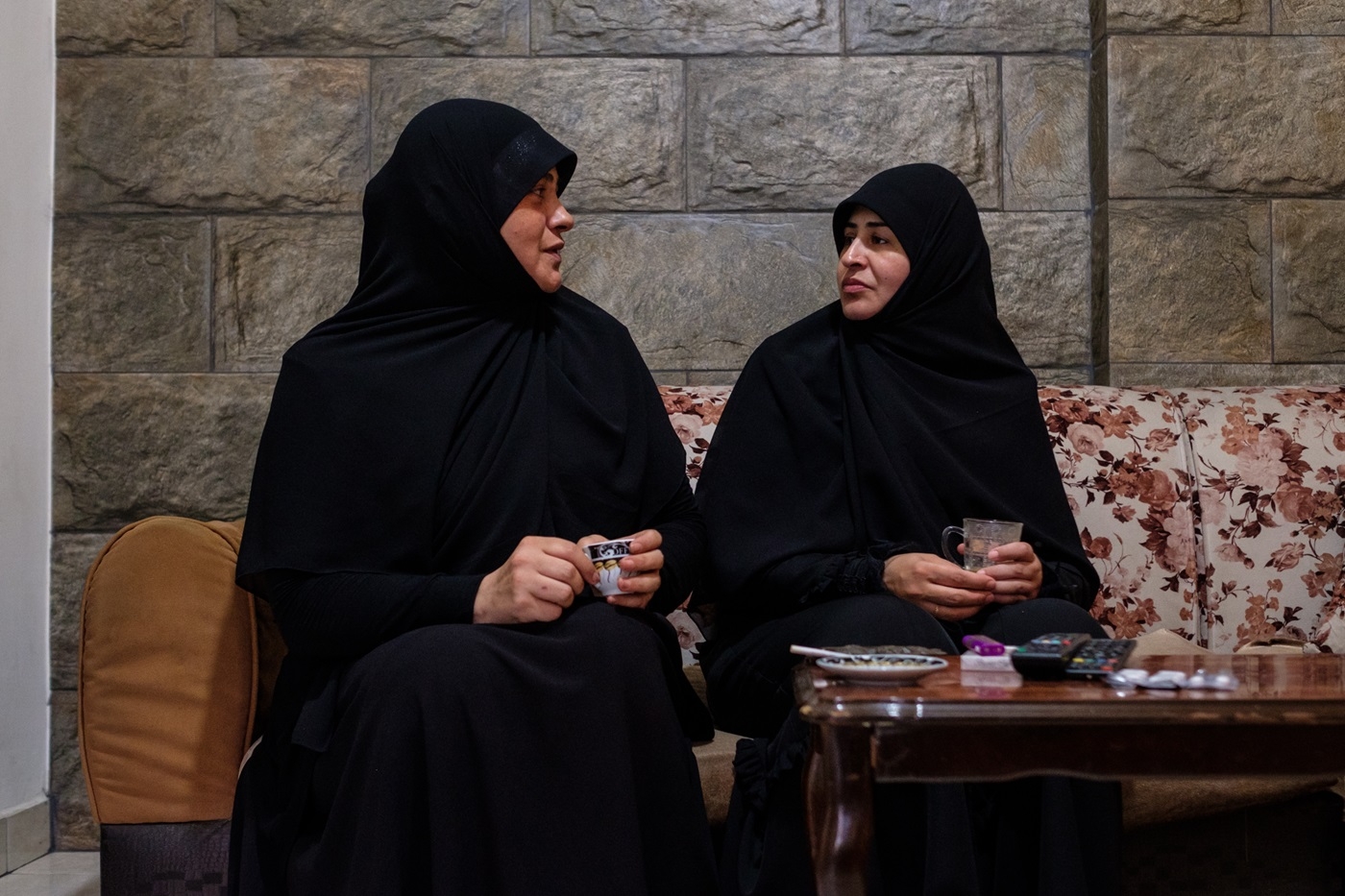 Two women holding small coffee cups and sitting side by side on a couch face each other in conversation.