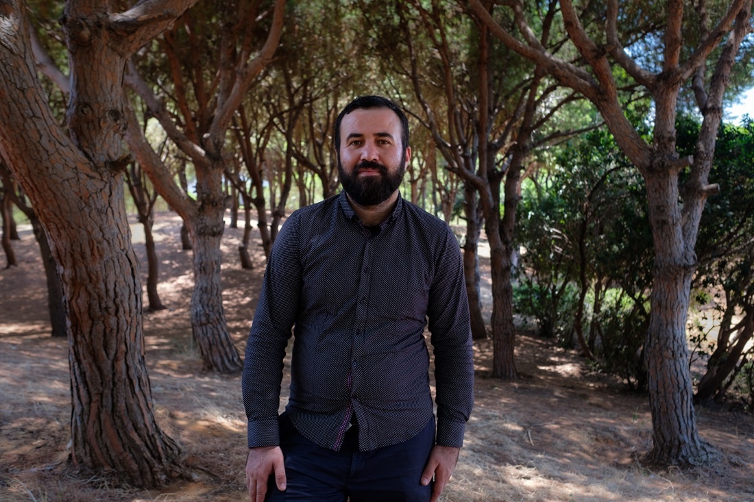 A smiling man stands among trees.