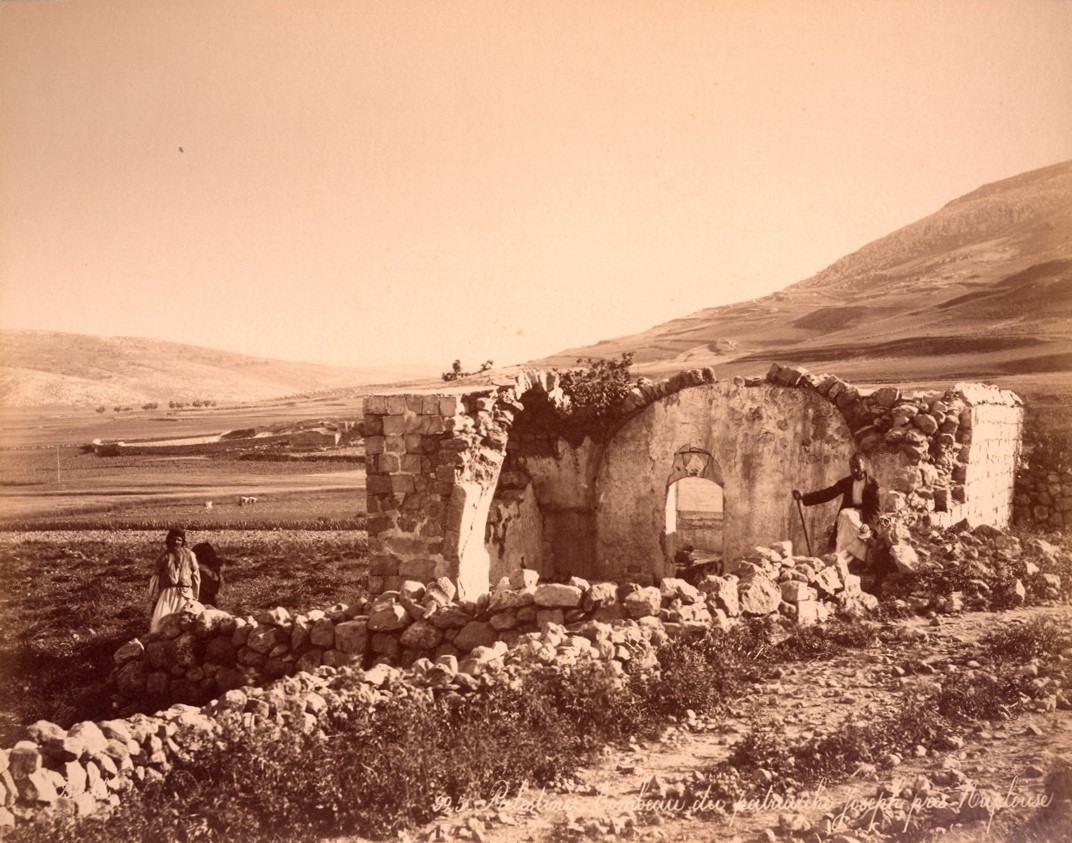 A sepia-toned photograph of a partially ruined stone structure in an open field with hills in the background, and two people standing near the structure.