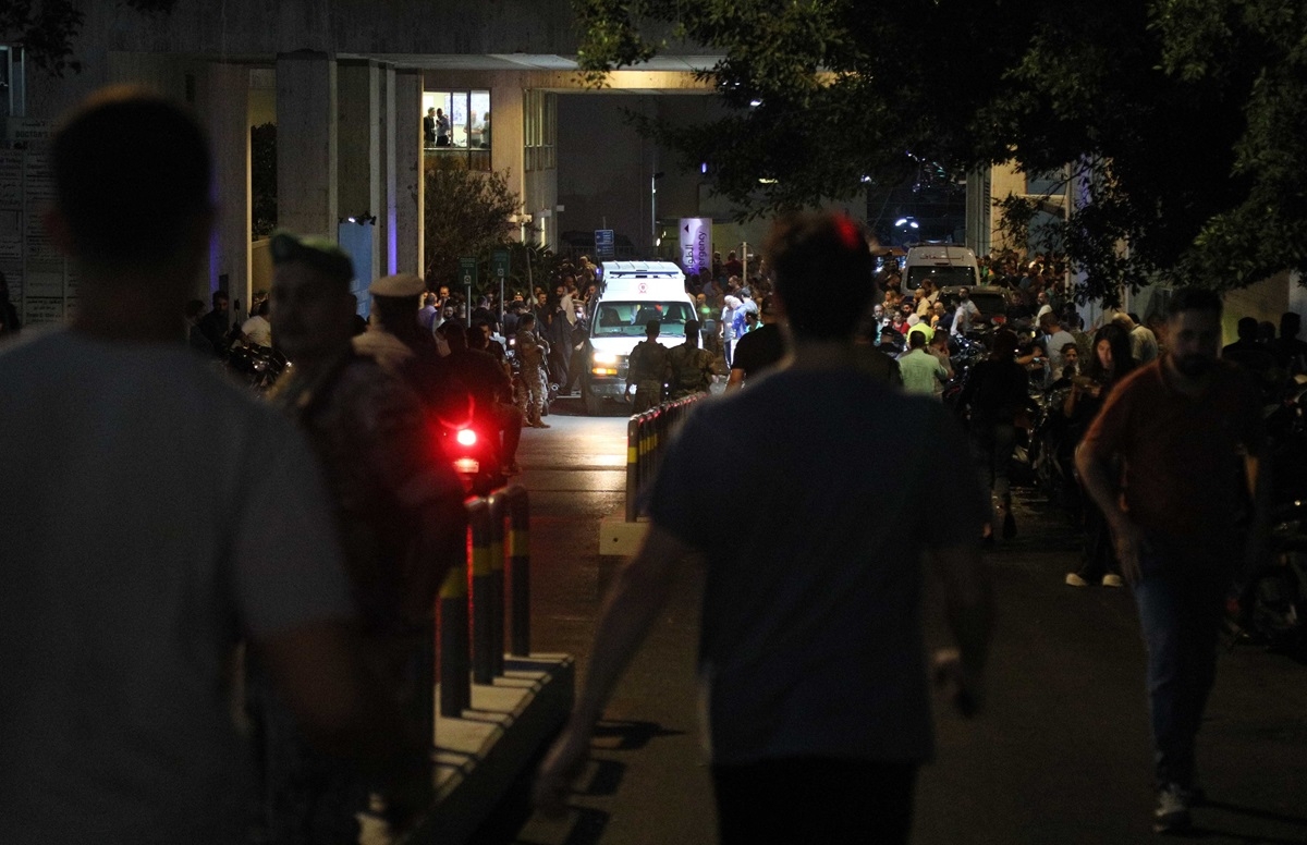 In the foreground are the the backs of men walking toward an ambulance arriving on a narrow street, surrounded by people.