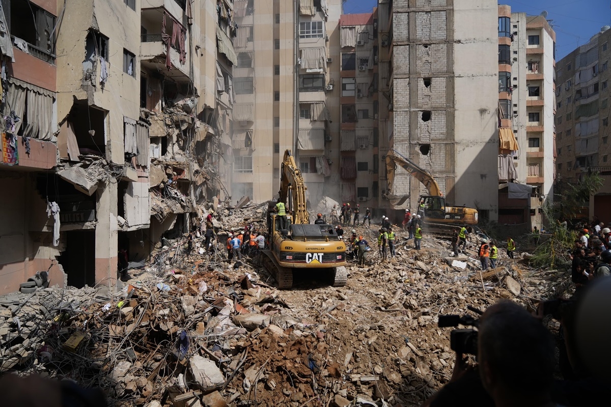 Two bulldozers and several people in high visibility vests stand atop a large mound of rubble, in the middle of a residential area, amid the destruction and ruin of nearby buildings.