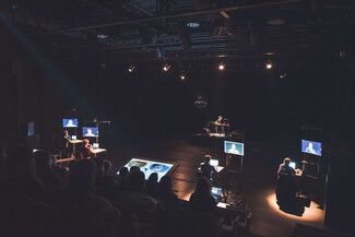 The audience watches five actors each sitting in front of a screen on stage.