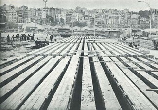 Black and white image of rows of vertically-arranged reinforced concrete piles with the Beirut skyline in the background and workers on the left side 