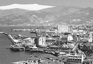 A black and white image of the port viewed from afar. Snowy Mount Sannine appears in the background, the silos are in the center of the image, and the rest of the port apparatus appears in the foreground.